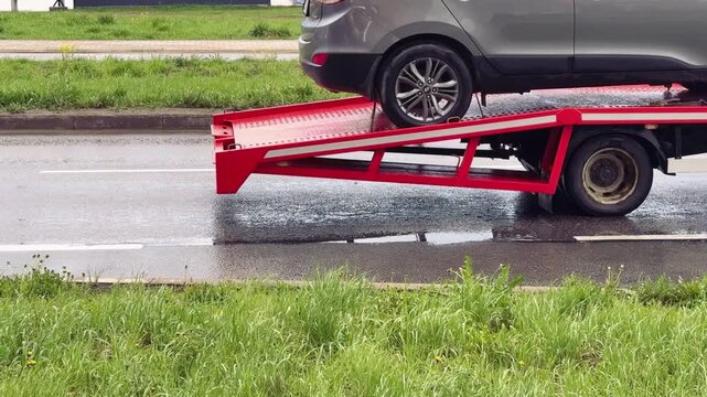 Close-up passenger car on red flatbed tow truck, spring roadside scene with wet pavement and puddles, green grassy verge dotted