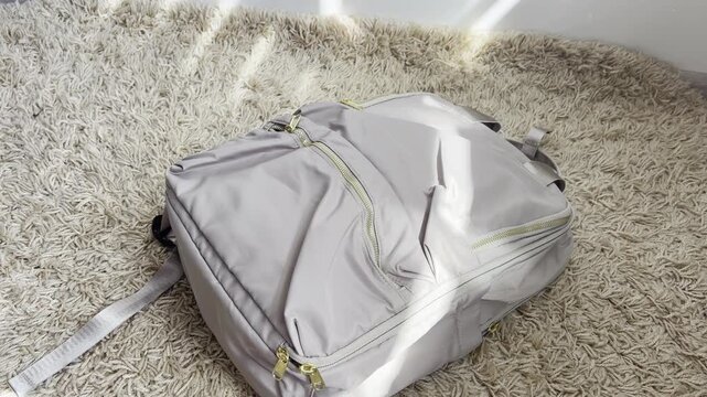 Casual gray backpack being dropped onto a fluffy carpet in a sunlit room with natural light streaming through a window creating dynamic shadows on the floor