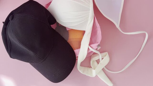 Top down view of travel preparation, showing a woman packing a white bikini top and a black baseball cap into a pink mesh organizer bag for a summer beach holiday