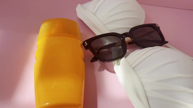 Summer vacation essentials featuring a white bikini, black sunglasses, and sunscreen bottle resting on a pink surface under a dynamic, shifting sunlight and shadows