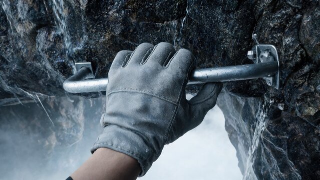 A gloved hand grips a metal climbing hold on a rocky surface. Mist surrounds the area, indicating a challenging climbing environment.