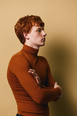 Young man with red hair wearing brown turtleneck sweater stands sideways with arms crossed against beige background. Profile portrait with serious expression and casual style. © SHOTPRIME STUDIO