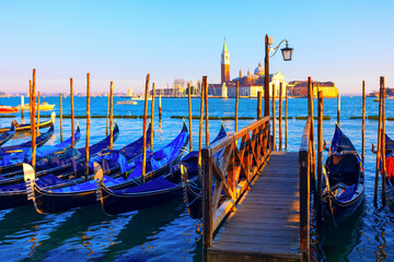Grand Canal in Venice, Italy, with blue gondolas moored along a wooden pier. Golden light of sunset illuminates the distant Church of San Giorgio Maggiore across the lagoon © Leilani