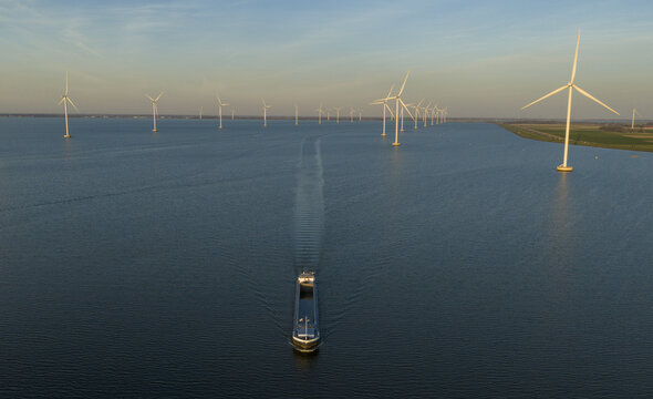 Aerial view of a large cargo ship sailing through an offshore wind farm with rows of turbines under a clear golden hour sky in Netherlands.