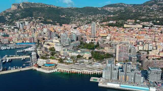 4K aerial view of Monte-Carlo Casino with green domes, Port Hercule superyacht marina, dense residential towers and T&ecirc;te de Chien rocky cliff above the principality of Monaco.