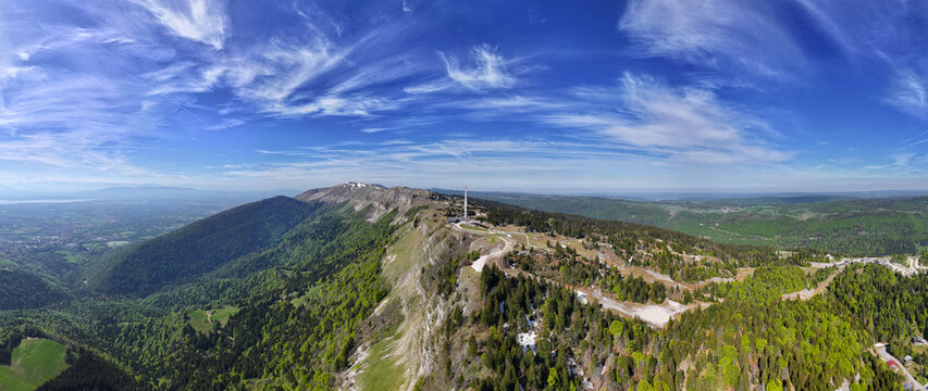 Aerial view of Col de la Faucille mountain pass featuring a telecommunications tower and lush green forests under a blue sky with wispy clouds in Gex, Auvergne-Rhone-Alpes, France.