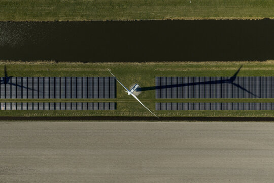 Aerial view of a wind turbine casting a long shadow over rows of solar panels next to a canal and agricultural fields in the Netherlands.