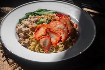 Fototapeta premium Traditional Thai-Chinese egg noodles with red BBQ pork, minced pork and green vegetables in a white bowl on a wooden table. Delicious Asian street food photography concept.