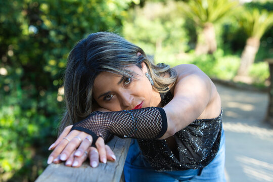 A young, beautiful woman, wearing make-up and dressed in jeans, gloves and a black T-shirt, leans her arms and face against the railing of a wooden bridge and gazes sensually at the camera.