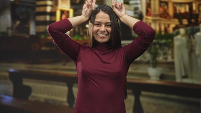 Woman smiling with fingers making bunny ears on her head in a maroon turtleneck inside a building church interior; playful.