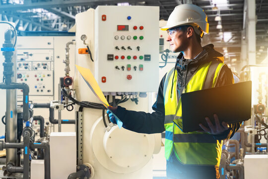 Industrial engineering, Plant maintenance. Engineer with laptop checks documents near control panel, pipes and production equipment.