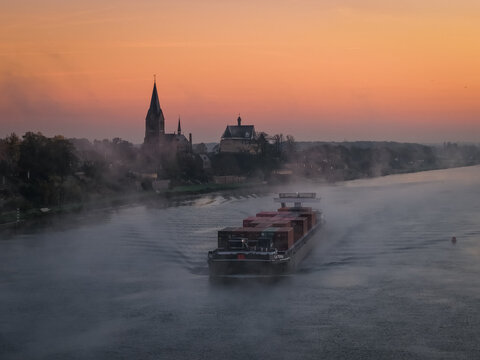Aerial view of a container barge navigating the misty Meuse river at sunrise with the silhouette of Kessel castle and church spire in the background Kessel, Limburg, Netherlands.