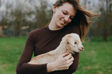 Naklejka premium Happy woman in a brown sweater holds a playful goat kid in a sunlit field, sharing a tender moment outdoors, showcasing gentle animal affection amid open nature and rural countryside