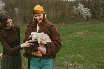 Naklejka premium Two women in casual coats share a moment outdoors as one holds a fluffy goat kid, smiling warmly, while the other offers a gentle touch on its head in a grassy field.