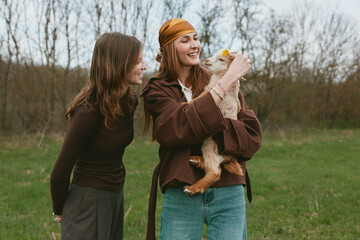 Naklejka premium Two women in casual clothes share a joyful moment with a goat kid in a grassy meadow, outdoor. A warm lifestyle scene highlighting bonding, care, and rural life.