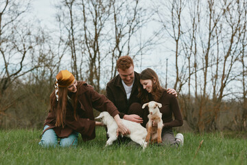 Fototapeta premium Three young adults enjoy a casual outdoor moment with two baby goats in a grassy field during early spring, capturing a warm and natural lifestyle scene.