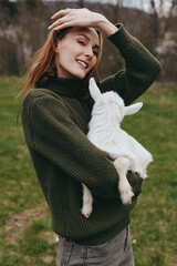 Naklejka premium woman goat kid outdoors lifestyle joyful smiling sweater field countryside sunlit nature affection cuddle animal companion rural grass meadow, a warm, candid moment captured with natural colors and