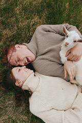 Naklejka premium Warm lifestyle scene with a smiling couple on the grass holding a goat kid, cozy sweaters, and natural light creating a tender, carefree mood in a peaceful outdoor setting
