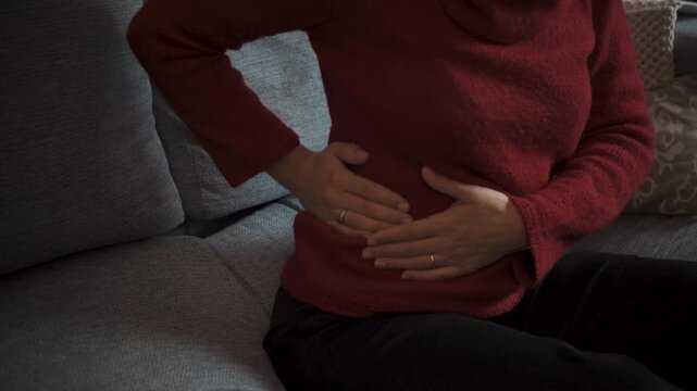 Young woman on her sofa at home suffering from lower back pain. Health and physical discomfort concept, video clip.