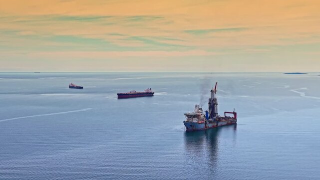 The oil tanker navigates the ocean waters with two other ships visible in the distance as the sun sets on the horizon creating a colorful sky.