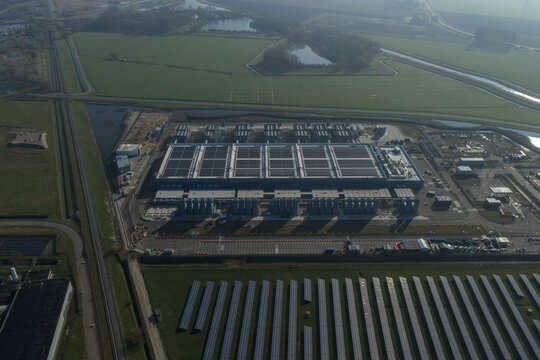 Aerial view of an AI data center with cooling towers and a cloud computing hub exterior featuring a blue industrial building, solar panels and green fields.