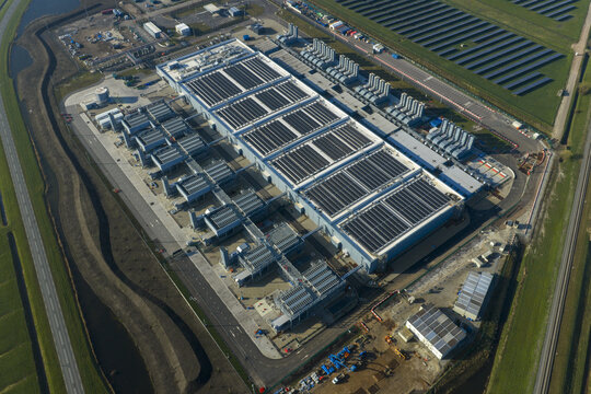 Aerial view of an AI data center and cloud computing hub exterior featuring rooftop cooling towers and solar panels surrounded by green fields.