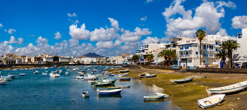 Arrecife Lanzarote showcasing colorful boats in the harbor set against a backdrop of historic buildings and volcanic mountains. The lively scene captures the island's vibrant coastal life