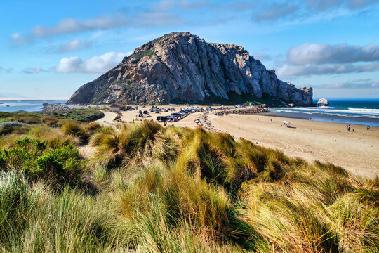 Morro Rock and beach area in Morro Bay, California, with visitors and parked cars near the shoreline. Popular coastal destination along the Pacific Coast with scenic views