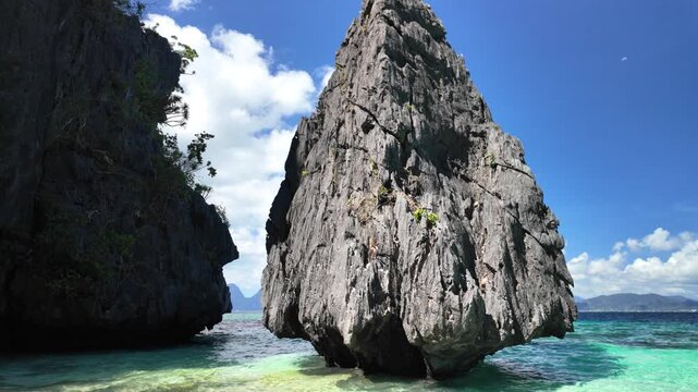 Entalula Beach featuring a dramatic limestone karst rock formation rising from the clear turquoise water, surrounded by tropical islands under a blue sky with clouds in El Nido, Palawan, Philippines