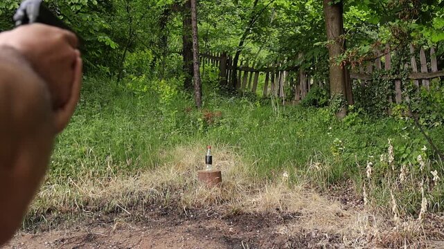 Focused bottle target with blurred hands aiming and cocking an airsoft pistol before firing outdoors, capturing shooting practice action in a natural rural environment.