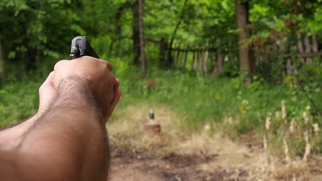 Close up of hands cocking and firing an airsoft pistol at a bottle target outdoors, showing bullet impact and the bottle falling after a successful shot in a natural environment.