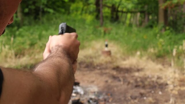 Over the shoulder view of a person aiming an airsoft pistol at a bottle outdoors, with visible shot impact and falling target after hit during realistic shooting practice in nature.