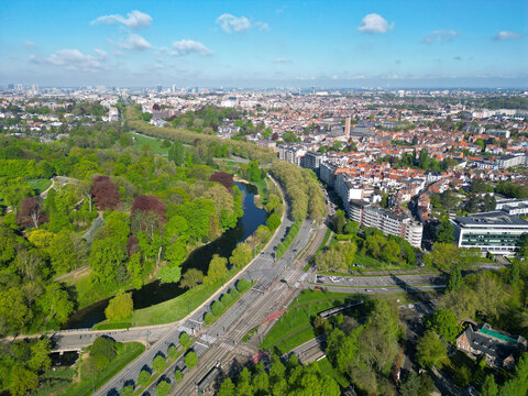 Aerial view of the lush green Parc de Woluwe with its winding lake, adjacent to urban residential blocks and a tram line under a bright blue sky in Woluwe-Saint-Pierre, Bruxelles, Belgium.