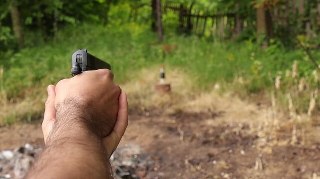 Close up shot of hands cocking and firing an airsoft pistol at a bottle target in a forest clearing, capturing the successful hit as the bottle falls after impact.