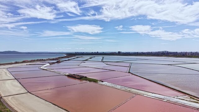 Saline ponds are visible in a coastal area. These ponds show various colors from the salt production process. Clear sky and distant city can be seen in the background.
