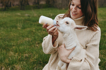 Naklejka premium A woman in a soft sweater gently bottles a hungry kid in an open field, capturing a tender moment of farm life, nurture, and animal care in a bright outdoor setting.