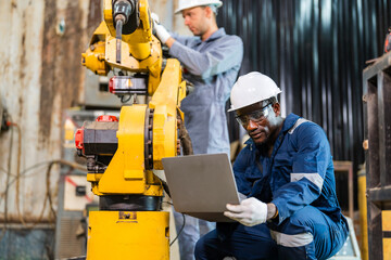 Two workers in hard hats operate machinery in an industrial workshop. One worker uses a laptop near a yellow robotic arm. The other worker stands behind the robotic arm © VStudio