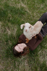 Naklejka premium A young woman lies on grass smiling as she holds a small white baby goat on her chest in a relaxed outdoor lifestyle scene.