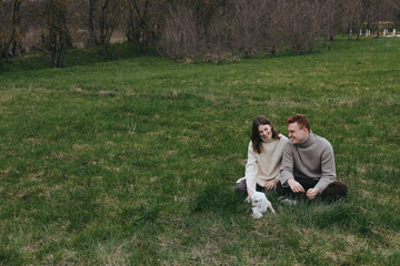 Naklejka premium Young couple sitting on green grass in casual sweaters enjoys spending time with a playful baby goat in a peaceful outdoor setting during early spring.