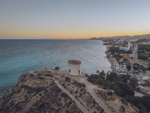 Aerial view of Santuari iber i roma de la Malladeta, a white circular tower on a rocky cliff overlooking the turquoise Mediterranean Sea at sunset in La Vila Joiosa, Comunidad Valenciana, Spain.