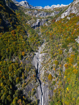 Aerial view of the Val di Mello waterfall cascading down steep granite cliffs surrounded by vibrant autumn foliage and jagged mountain peaks in Val Masino, Lombardia, Italy.
