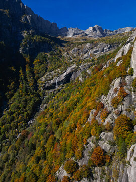 Aerial view of the Val di Mello valley featuring steep granite cliffs, autumn-colored forests, and a cascading waterfall under a clear blue sky in Val Masino, Lombardia, Italy.