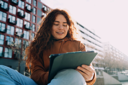 Happy red-haired student looking at digital tablet outdoors against city architecture. Remote education process, preparing for exams, online research, creative writing, digital nomad lifestyle.