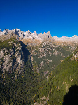Aerial view of the jagged granite peaks and dense evergreen forests of Val di Mello under a clear blue sky in Val Masino, Lombardia, Italy.