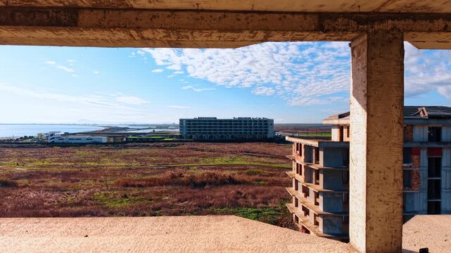 Sunny seashore with scarce greenery and buildings under construction. Flying inside the frame of the multi-storied structure of future hotel.