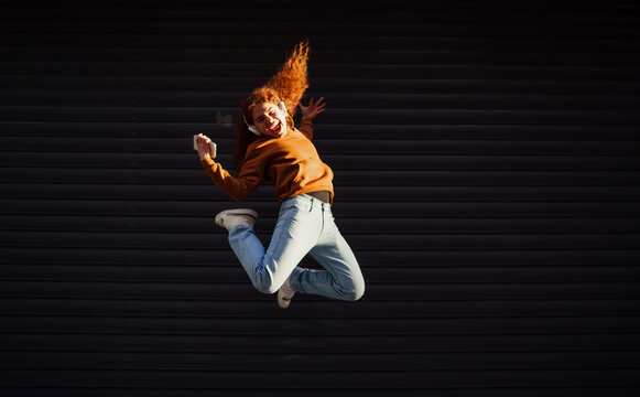 Cheerful redhead man with smartphone jumping high against dark wall, celebrating victory, success and achievement, active digital nomad, joy emotion, authentic Caucasian model person.