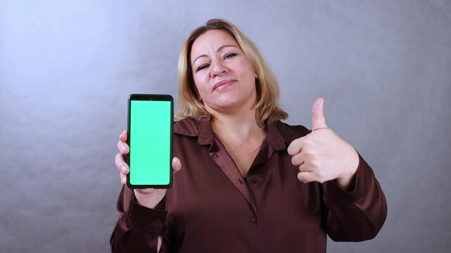 Caucasian woman with blonde hair displays smartphone with green screen while giving a thumbs up, showcasing positive engagement in a neutral studio setting