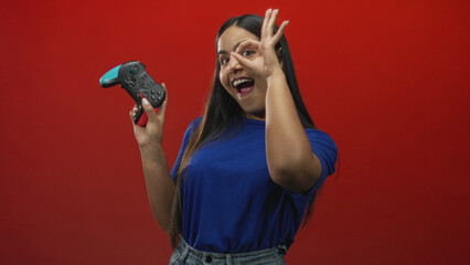 Woman in blue shirt holds gamepad and makes ok sign to eye against red backdrop in studio  playful confidence. © Krakenimages.com