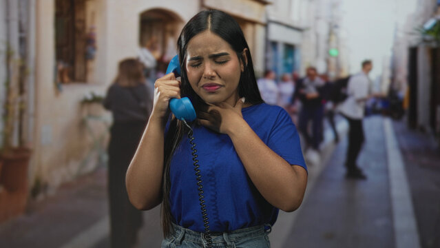 Young woman holding a blue telephone handset to her ear and clutching her neck on a busy urban street lined with storefronts and blurred pedestrians; panic plea.