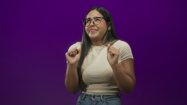 Young woman with glasses and hands raised, recoiling with a disgusted facial grimace, wearing beige tee and jeans in a purple studio; disgust avoidance.